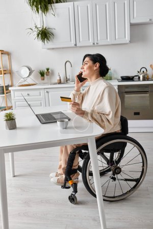 Foto de Una mujer discapacitada en silla de ruedas hablando en un teléfono celular mientras trabaja de forma remota desde su cocina. - Imagen libre de derechos
