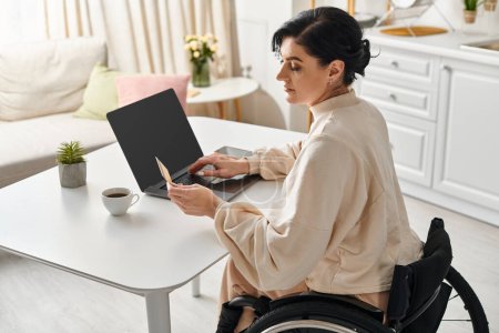 Una mujer en silla de ruedas está utilizando un ordenador portátil en su cocina, dedicándose a un trabajo remoto.