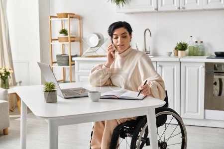 Une femme en fauteuil roulant travaille à distance à une table avec un ordinateur portable dans sa cuisine.