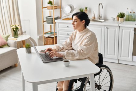 A disabled woman in a wheelchair works remotely on her laptop in the kitchen, showcasing digital independence.