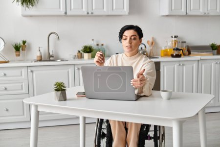Una mujer discapacitada en silla de ruedas está enfocada en su computadora portátil en una mesa de cocina, participando en actividades de trabajo o de ocio remotas..