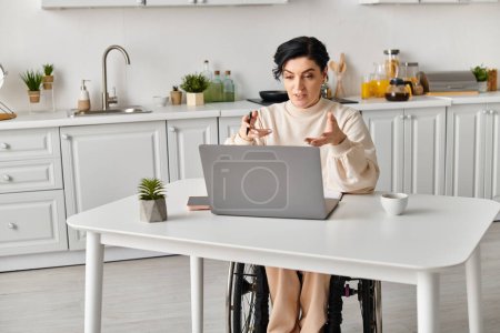 Une femme handicapée dans un fauteuil roulant assise à une table de cuisine, concentrée sur son ordinateur portable. Elle travaille à distance..