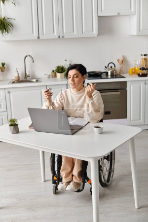 A disabled woman in a wheelchair works remotely in her kitchen, focusing on a laptop computer on the table.