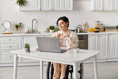 Una mujer en silla de ruedas trabaja de forma remota en una mesa de cocina, usando una computadora portátil para mantenerse conectada y productiva.