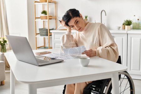 Une femme handicapée dans un fauteuil roulant travaille sur son ordinateur portable à une table dans sa cuisine.