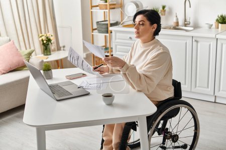 Une femme handicapée dans un fauteuil roulant travaille sur un ordinateur portable à une table dans sa cuisine.