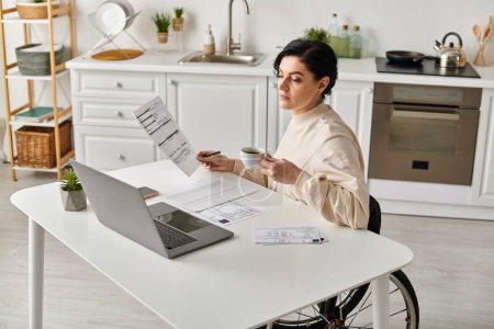 A woman in a wheelchair works on her laptop, surrounded by papers, in a cozy kitchen setting.
