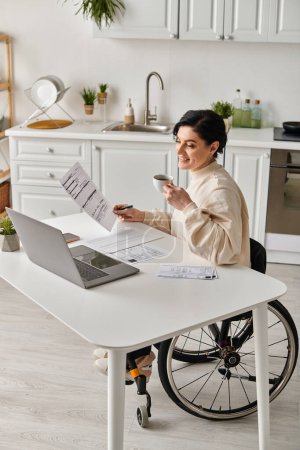 Una mujer en silla de ruedas está sentada en una mesa en su cocina, enfocada en trabajar en su computadora portátil remotamente.