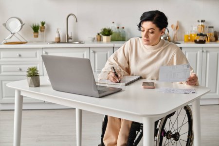 Une femme handicapée dans un fauteuil roulant assise à une table avec un ordinateur portable, travaillant à distance dans sa cuisine.