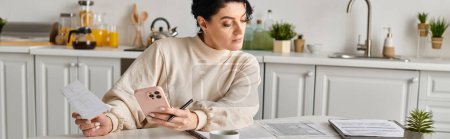 Photo for Woman using a cellphone while working remotely from her kitchen table. - Royalty Free Image