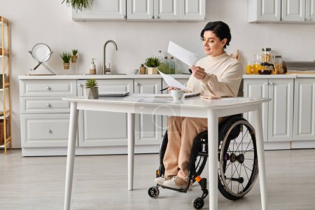 Une femme en fauteuil roulant s'assoit dans sa cuisine, profondément réfléchie alors qu'elle tient un bout de papier, concentrée sur son travail créatif.