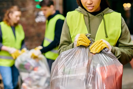 Foto de A young woman in gloves and safety vest holding a bag of garbage, recycling with fellow volunteers. - Imagen libre de derechos
