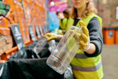 Foto de A woman in a yellow vest holds a plastic cup while sorting trash with other eco-conscious volunteers. - Imagen libre de derechos