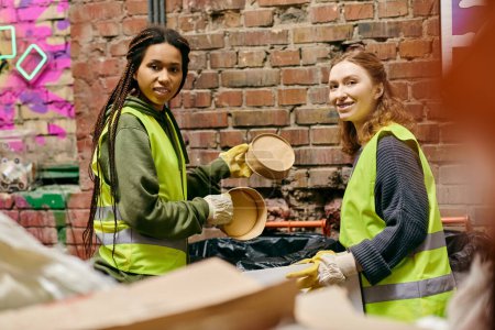Photo for Two young female volunteers in safety vests and gloves sorting through trash together. - Royalty Free Image