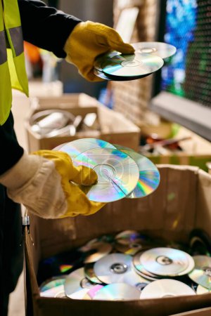 Téléchargez les photos : Young volunteer in yellow safety vest carefully holding a CD while sorting waste, emphasizing eco-conscious behavior. - en image libre de droit