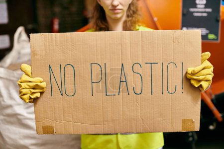 Young volunteer in gloves and safety vest advocates against plastic waste by holding a no plastic sign.