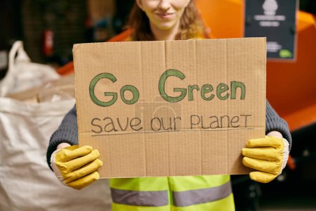 A young volunteer in gloves and safety vest holds a sign that says go green save our planet in a passionate call to action.