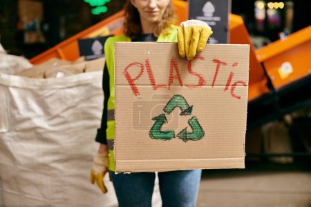 Foto de A young volunteer in gloves and a safety vest holding a cardboard sign that says Plastic while sorting waste. - Imagen libre de derechos