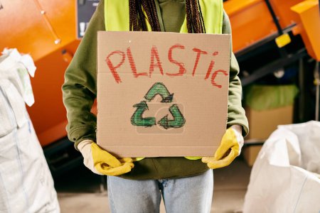 A young volunteer in gloves and safety vest holding a cardboard sign that says plastic.