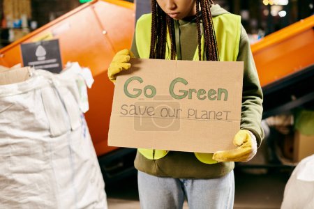 A young volunteer in gloves and a safety vest holds a sign urging to go green and save our planet.