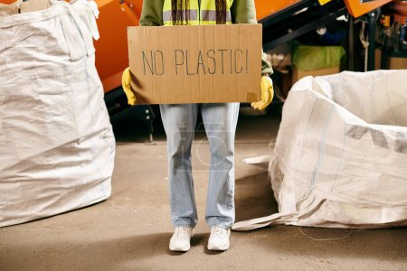 Young volunteer in gloves and safety vest holds sign that says no plastic while sorting waste.