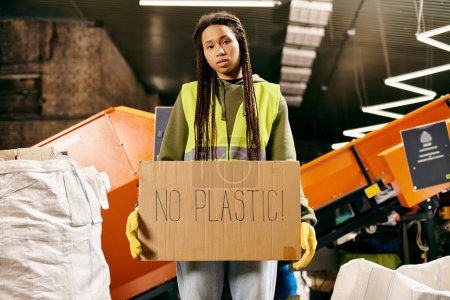 Young volunteer in gloves and safety vest holding a sign that says no plastic while sorting waste.
