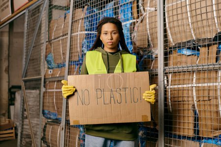 Foto de A young african american volunteer in gloves and safety vest holds a sign that says no plastic while sorting waste. - Imagen libre de derechos