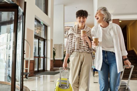 Téléchargez les photos : Deux lesbiennes âgées se promènent joyeusement dans un couloir de l'hôtel avec leurs bagages. - en image libre de droit