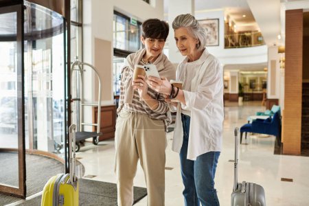 Two women, a loving senior lesbian couple, standing in a lobby engrossed in a cell phone.