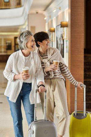 Two senior lesbian women happily walk down a hotel hallway with luggage.