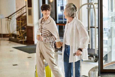 Photo for Loving senior lesbian couple standing with luggage in elegant hotel lobby. - Royalty Free Image