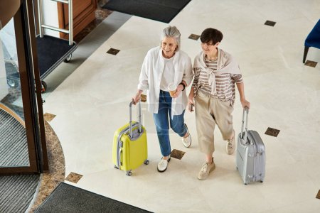 Loving senior lesbian couple strolling down hotel hallway with luggage.