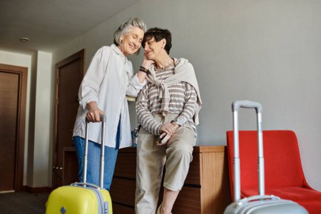 A senior lesbian couple standing together with a suitcase in a hotel.