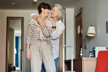 Senior lesbian couple stand in a cozy living room, sharing a moment of intimacy and connection.