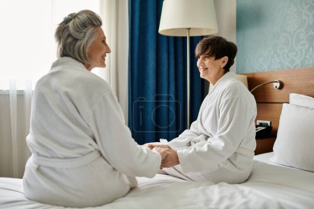 Senior lesbian couple sitting together on a bed, wearing robes, sharing a tender moment.