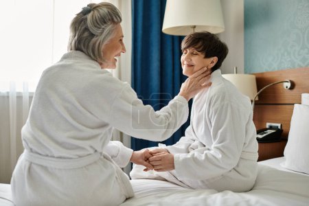 Photo for Senior lesbians embrace on bed, one in white robe, showing loving tenderness. - Royalty Free Image