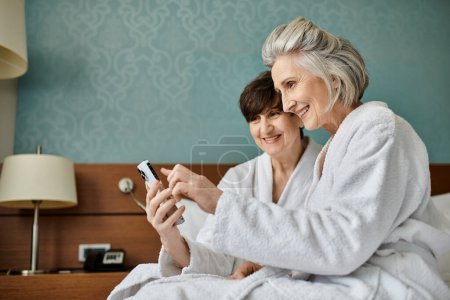 Couple lesbien sénior dans une étreinte aimante sur un lit, absorbé dans l'écran d'un téléphone portable.