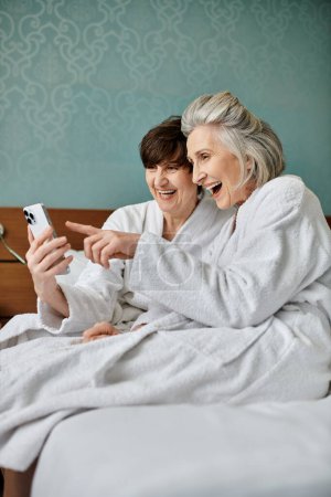 Zarte ältere lesbische Paar und junge Frau genießen Zeit zusammen auf einem Bett mit einem Mobiltelefon.