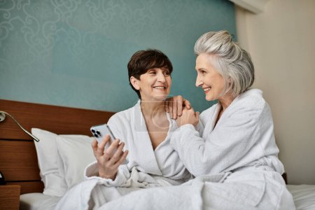 Ein älteres lesbisches Paar in Roben sitzt eng auf einem Bett und zeigt Liebe und Zärtlichkeit.