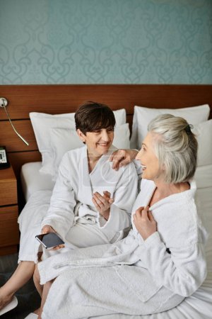 Senior lesbian couple on a bed in robes, sharing a tender moment.