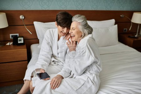 Affectionate senior lesbian couple sitting on top of hotel bed, sharing a loving moment.