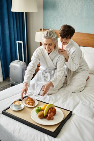 Senior lesbian couple enjoying a cozy meal on a bed.