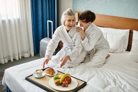 Senior lesbian couple in white robes on bed, showing love and tenderness.