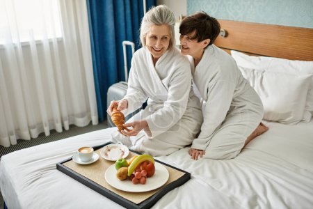 Ältere lesbische Paar genießen einen zarten Moment, sitzt auf einem gemütlichen Bett in einem Hotelzimmer.