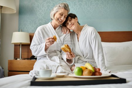 Foto de Una pareja lesbiana mayor con túnicas, sentada en una cama, exudando ternura y amor. - Imagen libre de derechos