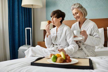 Senior lesbian couple serenely sitting on a cozy bed in a hotel room.