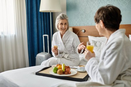 Senior lesbiana pareja en albornoces disfruta de una bandeja de fruta en la cama.