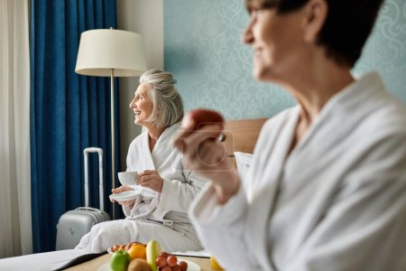 Foto de Dos mujeres mayores en un abrazo amoroso, sentadas juntas encima de una cama en una habitación de hotel. - Imagen libre de derechos