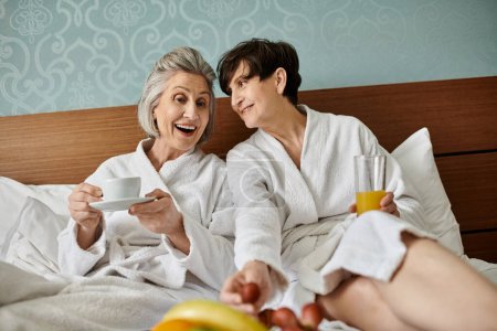 Two women in white robes, a senior lesbian couple, sit peacefully on a bed.