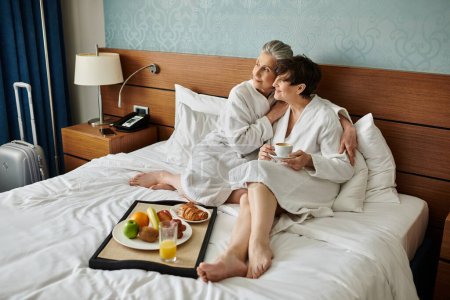 Senior lesbian couple sitting tenderly on top of a cozy bed.
